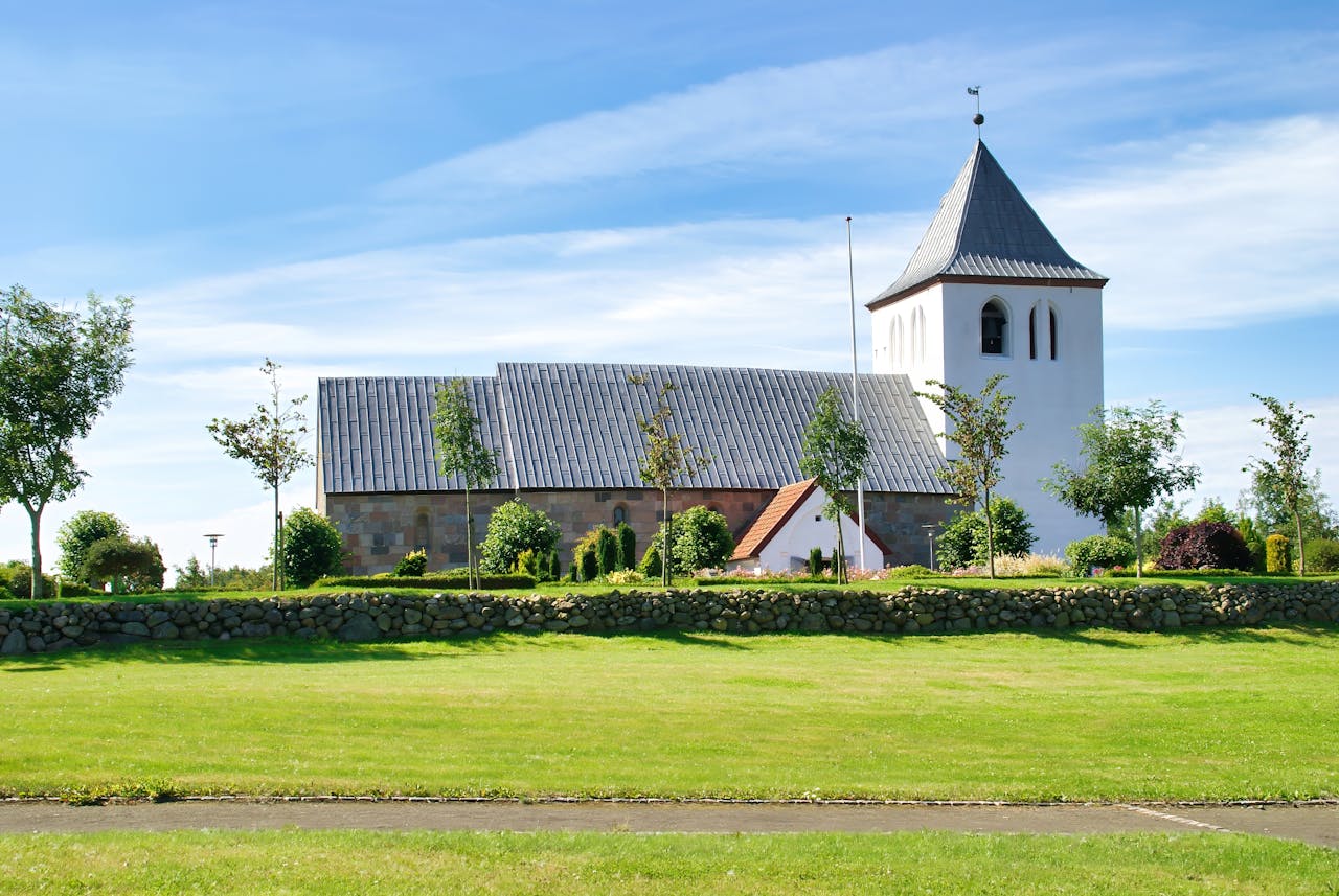 home-hero Beautiful view of Mabjerg Church in Denmark, surrounded by lush greenery and clear blue skies.